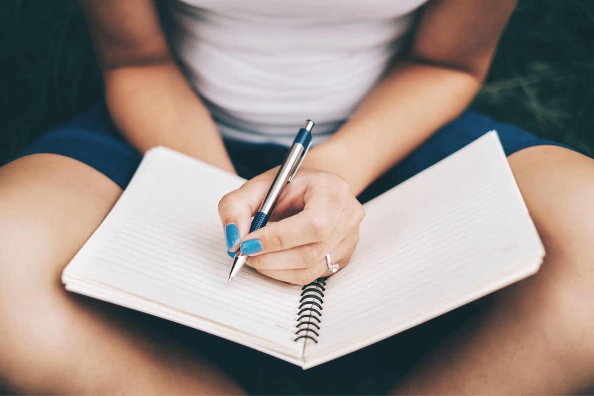 image of woman with engagement ring, sitting crosslegged with a notebook, writing